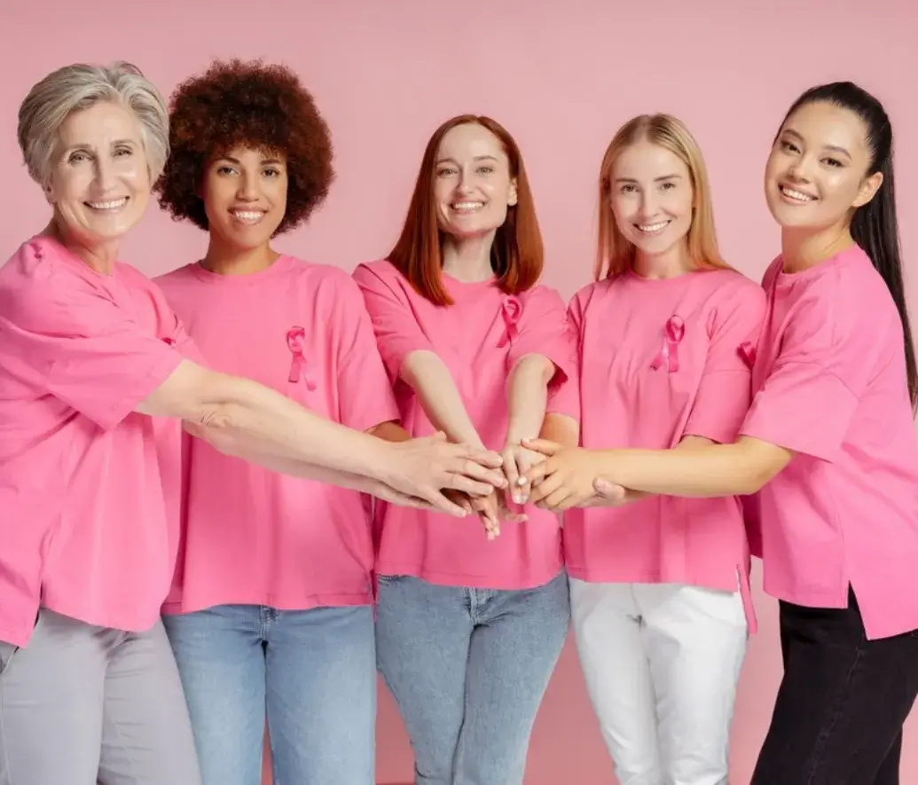 Group of diverse women smiling and holding hands, united in support of Breast Cancer Awareness Month, all wearing pink t-shirts with ribbons.
